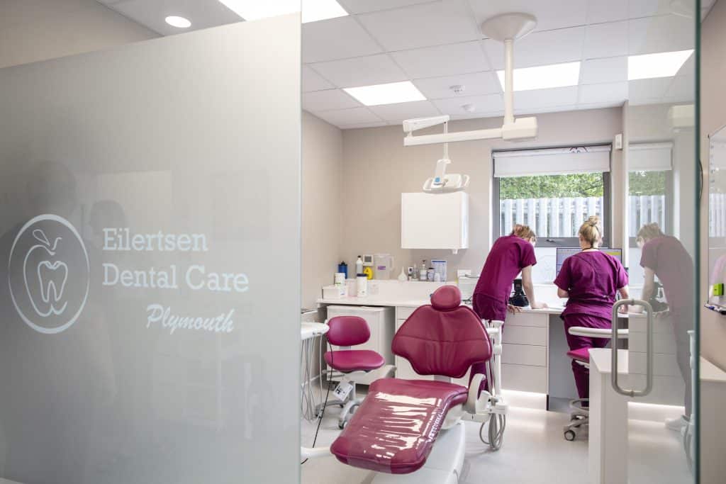 Modern treatment room at Eilertsen Dental Care Plymouth with a burgundy dental chair, clinical equipment, and team members preparing at a workstation.